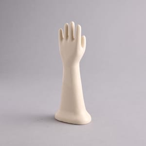 May include: A smooth, off-white ceramic hand display stand featuring an upright, open-palm posture with fingers slightly spread. The structure includes a long, tapered wrist section that extends down to a stable, rounded base for upright positioning.