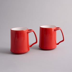 May include: Two cylindrical ceramic mugs with a vibrant red exterior and a smooth white interior. Each mug features a distinctively angular, geometric handle attached to the side, providing a modern silhouette for these beverage containers.