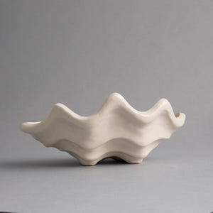 May include: A cream-colored ceramic bowl with a distinct wavy, scalloped rim that mimics the organic shape of a seashell. The bowl features a smooth, glossy finish and a deep, curved basin, resting on a base that follows the undulating form.