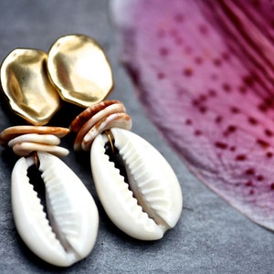 Op de afbeelding: Gouden oorbellen met een gelaagd schelpdesign en een witte kauri schelp hanger.