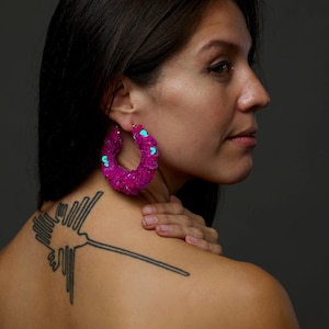 Op de afbeelding: Een vrouw met lang donker haar draagt grote roze oorringen met turquoise accenten. Ze heeft een tatoeage van een gestileerde vogel op haar rug.