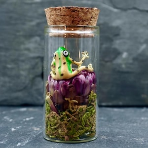 Puede incluir: Una pequeña botella de vidrio con tapón de corcho, que contiene una figura de rana. La rana es verde y amarilla, sentada sobre una flor morada y musgo. La botella está sobre una superficie gris oscura.
