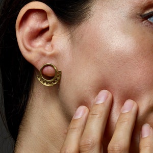 May include: A close-up of a person's ear with a gold, geometric, half-circle earring. The earring has a textured surface and is worn in the left ear.