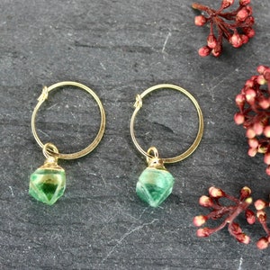 Puede incluir: Un par de pendientes de aro dorados con colgantes de piedras preciosas verdes facetadas. Los pendientes se exhiben sobre una superficie gris oscuro, con ramitas de bayas rojas en el fondo. La joyer&iacute;a es elegante y adecuada para el uso diario.