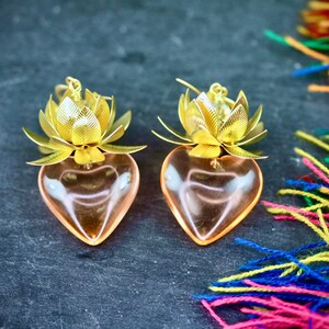 以下が含まれることがあります： 蓮の花のデザインが施された、金色のハート型のイヤリングのペア。イヤリングは透明なガラス製で、ピンクがかった色合いをしています。
