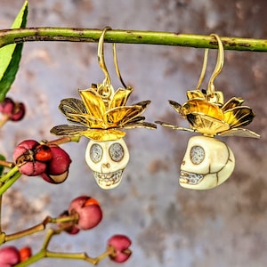 May include: A pair of gold-colored earrings featuring a skull design. The skulls are cream-colored with silver accents and are topped with a gold flower. The earrings hang from gold hooks and are displayed against a blurred background.