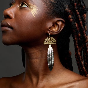 Op de afbeelding: Een vrouw met gevlochten haar draagt ​​een paar gouden oorbellen met een zon- en veertjesontwerp. De oorbellen zijn lang en hangen aan haar oorlel.