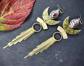 May include: A pair of long, gold earrings with a crescent moon shape and a red and white patterned bead. The earrings have a fringe of gold strands hanging from the bottom.