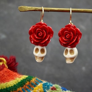 May include: A pair of dangle earrings with a white skull charm topped with a red rose. The earrings are hanging from a gold stand.