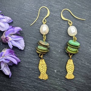 Può includere: Orecchini color oro con un ciondolo a forma di pesce, con una perla bianca, perline verdi impilate e un ciondolo a forma di pesce. Gli orecchini sono esposti accanto a fiori viola su una superficie scura.