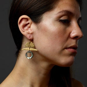 May include: Gold-toned earrings with a geometric design. The earrings feature a sunburst shape at the top, a horizontal bar, and a circular element with iridescent accents. The earrings are suspended from a gold hook.