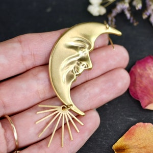 May include: Gold crescent moon earring with a face sculpted into the moon. The earring has a sunburst design at the bottom.