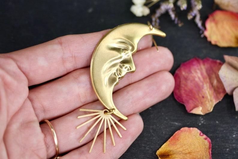 May include: Gold crescent moon earring with a face sculpted into the moon. The earring has a sunburst design at the bottom.