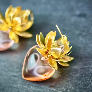 以下が含まれることがあります： ピンクのハート型のペンダントと蓮の花のデザインが施された、ゴールドトーンのイヤリングのペアです。