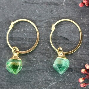 Puede incluir: Un par de pendientes de aro dorados con un colgante de piedra verde facetada. Los pendientes se muestran sobre una superficie oscura y texturizada, con una ramita de bayas rojas. Joyas.