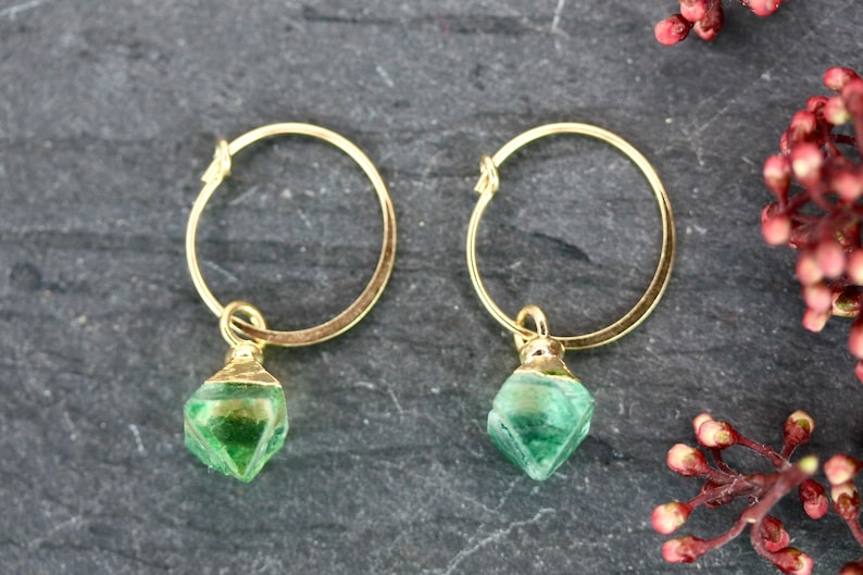 Puede incluir: Un par de pendientes de aro dorados con un colgante de piedra verde facetada. Los pendientes se muestran sobre una superficie oscura y texturizada, con una ramita de bayas rojas. Joyas.
