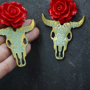 Puede incluir: Un par de pendientes de calavera de toro dorados con detalles de rosas rojas. Las calaveras de toro est&aacute;n detalladas con un patr&oacute;n floral.