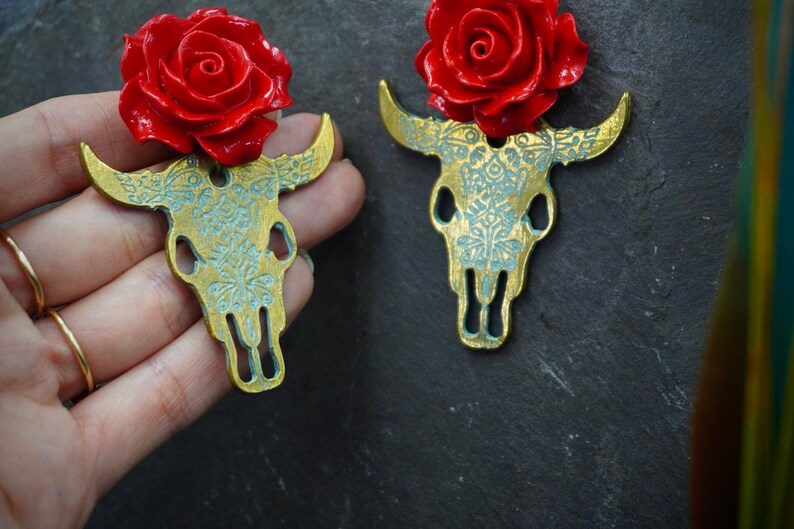 Puede incluir: Un par de pendientes de calavera de toro dorados con detalles de rosas rojas. Las calaveras de toro est&aacute;n detalladas con un patr&oacute;n floral.