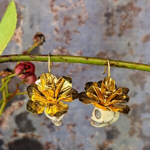 May include: A pair of gold-colored earrings featuring a skull design beneath a gold flower. The earrings are suspended from gold hooks. The skulls are off-white with detailed facial features. The background is a muted blue and gray.