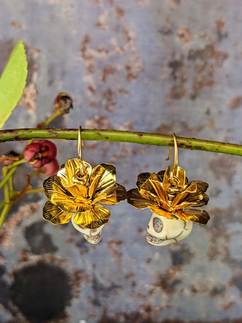 May include: A pair of gold-colored earrings featuring a skull design beneath a gold flower. The earrings are suspended from gold hooks. The skulls are off-white with detailed facial features. The background is a muted blue and gray.