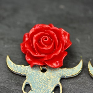 Puede incluir: Un par de pendientes con un encanto de rosa roja en la parte superior de un dise&ntilde;o de cr&aacute;neo de toro azul y dorado.