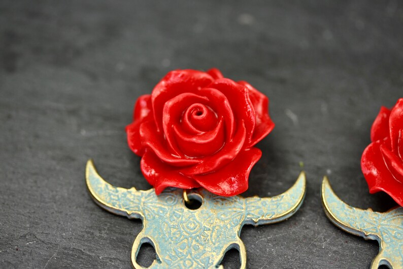Puede incluir: Un par de pendientes con un encanto de rosa roja en la parte superior de un dise&ntilde;o de cr&aacute;neo de toro azul y dorado.