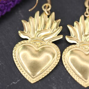 K&ouml;nnte beinhalten: Ein Paar goldfarbene Ohrringe mit einem Herz mit Flammenmotiv. Das Herz ist mit einem Perlenrand verziert.
