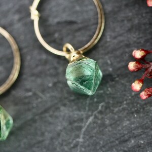 Puede incluir: Primer plano de dos pendientes de aro dorados, cada uno con un colgante de piedra preciosa verde facetada. Los pendientes est&aacute;n sobre un fondo oscuro y texturizado, con una ramita de capullos rojos en la esquina superior derecha.