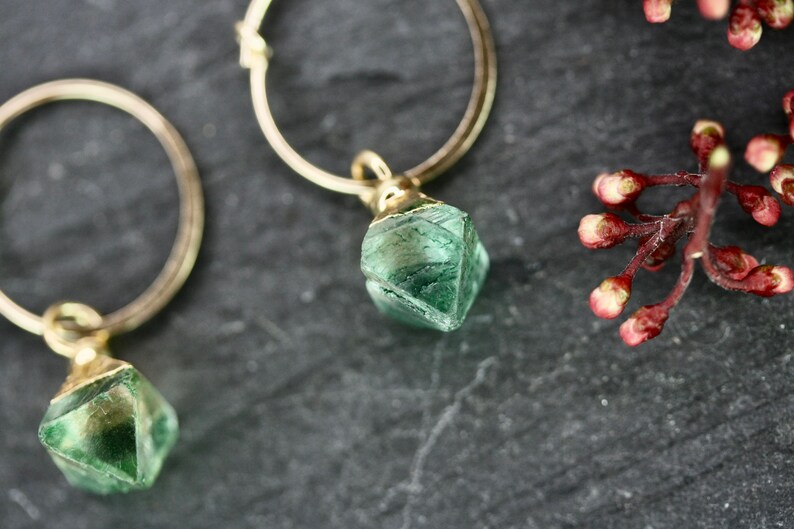 Puede incluir: Primer plano de dos pendientes de aro dorados, cada uno con un colgante de piedra preciosa verde facetada. Los pendientes est&aacute;n sobre un fondo oscuro y texturizado, con una ramita de capullos rojos en la esquina superior derecha.