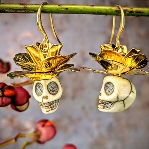 May include: Gold-toned earrings featuring a skull design with a flower accent. The skull has sparkling eyes and a detailed jawline. The earrings are suspended from gold-colored hooks and are set against a blurred background.