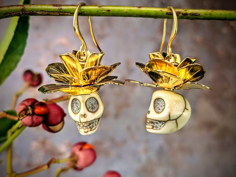 May include: Gold-toned earrings featuring a skull design with a flower accent. The skull has sparkling eyes and a detailed jawline. The earrings are suspended from gold-colored hooks and are set against a blurred background.