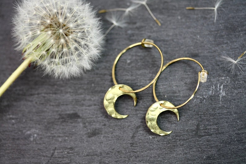 May include: Gold-colored hoop earrings with hammered crescent moon charms. The earrings are displayed on a dark gray surface, with a dandelion and scattered seeds in the background. The earrings have a simple, elegant design.