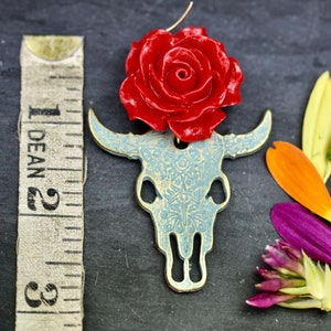 Puede incluir: Un amuleto de rosa roja con un colgante de calavera de toro con estampado azul y blanco. El amuleto est&aacute; hecho de metal y tiene un aspecto vintage.