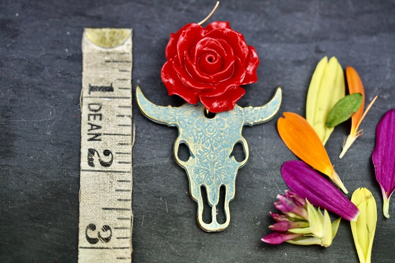 Puede incluir: Un amuleto de rosa roja con un colgante de calavera de toro con estampado azul y blanco. El amuleto est&aacute; hecho de metal y tiene un aspecto vintage.