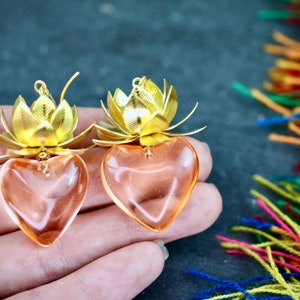 以下が含まれることがあります： 蓮の花のデザインが施された、ゴールドとピンクのハート型のイヤリングのペア。イヤリングは金属とガラスでできています。