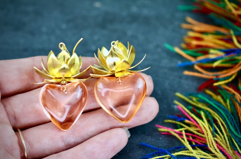 以下が含まれることがあります： 蓮の花のデザインが施された、ゴールドとピンクのハート型のイヤリングのペア。イヤリングは金属とガラスでできています。