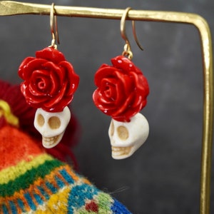 May include: A pair of dangle earrings with a white skull and a red rose on top. The earrings are hanging from a gold stand.