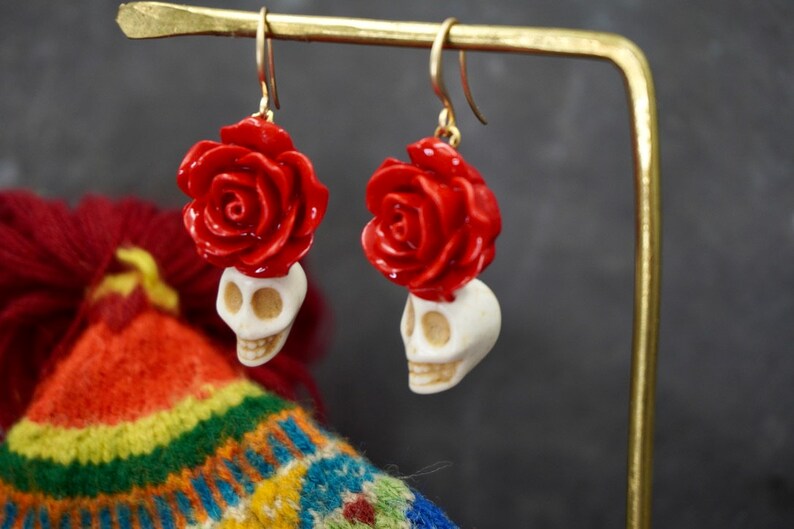 May include: A pair of dangle earrings with a white skull and a red rose on top. The earrings are hanging from a gold stand.