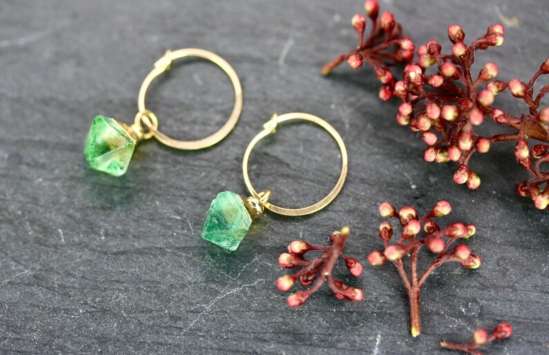 Puede incluir: Dos pendientes de aro dorados con colgantes de piedras preciosas facetadas de color verde claro. Los pendientes se exhiben sobre una superficie gris oscura, con ramitas de capullos rojos y rosas. Una joya delicada.