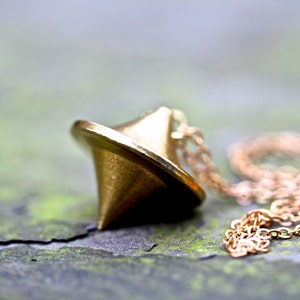 Op de afbeelding: Een gouden draaiend top-hanger aan een gouden ketting. De hanger is een eenvoudig, geometrisch ontwerp met een puntige punt.