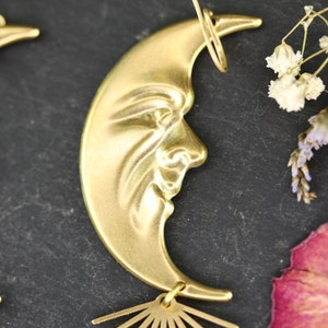May include: A pair of gold crescent moon earrings with a face design and a sunburst dangle. The earrings are on a dark gray background.
