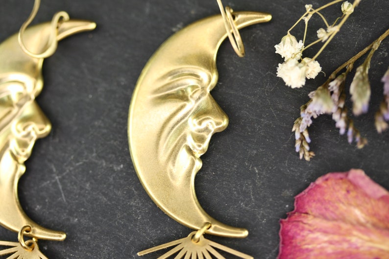 May include: A pair of gold crescent moon earrings with a face design and a sunburst dangle. The earrings are on a dark gray background.