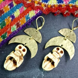 May include: A pair of gold tone earrings with a skull charm and a crescent moon design. The earrings are hanging from a woven fabric background.