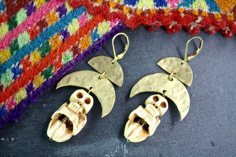 May include: A pair of gold tone earrings with a skull charm and a crescent moon design. The earrings are hanging from a woven fabric background.