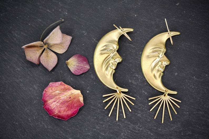 May include: A pair of gold crescent moon earrings with a face design and a sunburst dangle. The earrings are on a dark gray background.