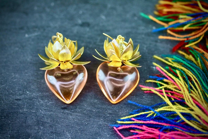 以下が含まれることがあります： 蓮の花のデザインが施された、金色のハート型のイヤリングのペア。イヤリングは透明なガラス製で、ピンクがかった色合いをしています。