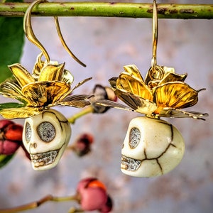 May include: Gold-colored earrings featuring a skull design with a flower accent. The skull is off-white with dark cracks and sparkling eyes and teeth. The flower is gold with detailed petals. The earrings hang from gold hooks.