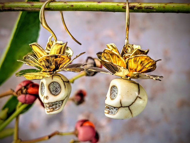May include: Gold-colored earrings featuring a skull design with a flower accent. The skull is off-white with dark cracks and sparkling eyes and teeth. The flower is gold with detailed petals. The earrings hang from gold hooks.