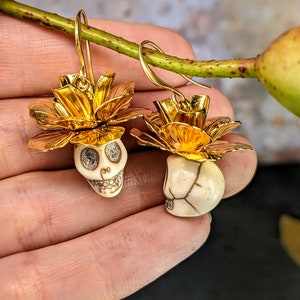 May include: A pair of gold-colored earrings featuring skull-shaped pendants. The skulls are white with detailed facial features and are topped with gold-colored flower-like accents. The earrings have a hook closure.