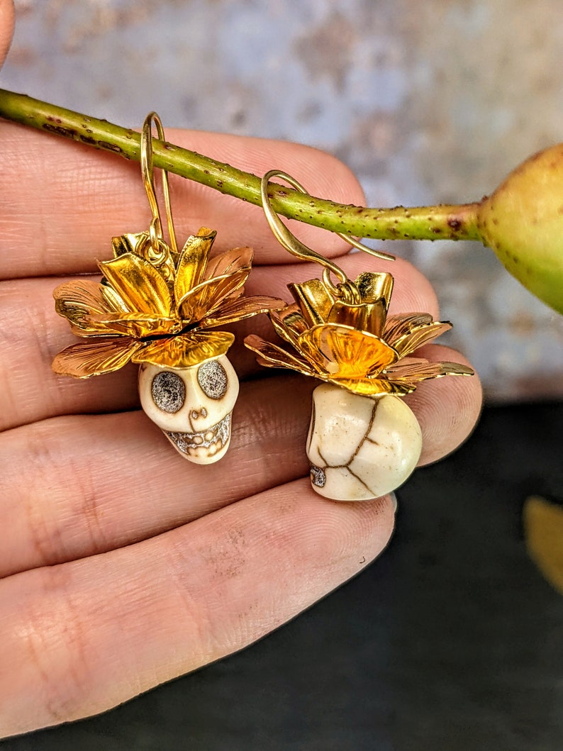 May include: A pair of gold-colored earrings featuring skull-shaped pendants. The skulls are white with detailed facial features and are topped with gold-colored flower-like accents. The earrings have a hook closure.